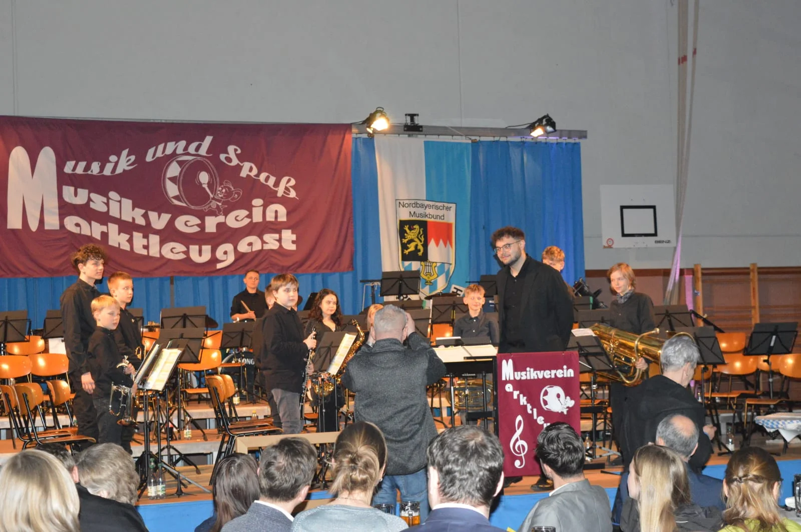 Die Nachwuchsmusiker stehen auf der Bühne und es wird von den Zuhörern applaudiert. Ein Fotograf steht vor der Bühne. Der Dirigent Jonas Trapper lächelt in die Kamera.