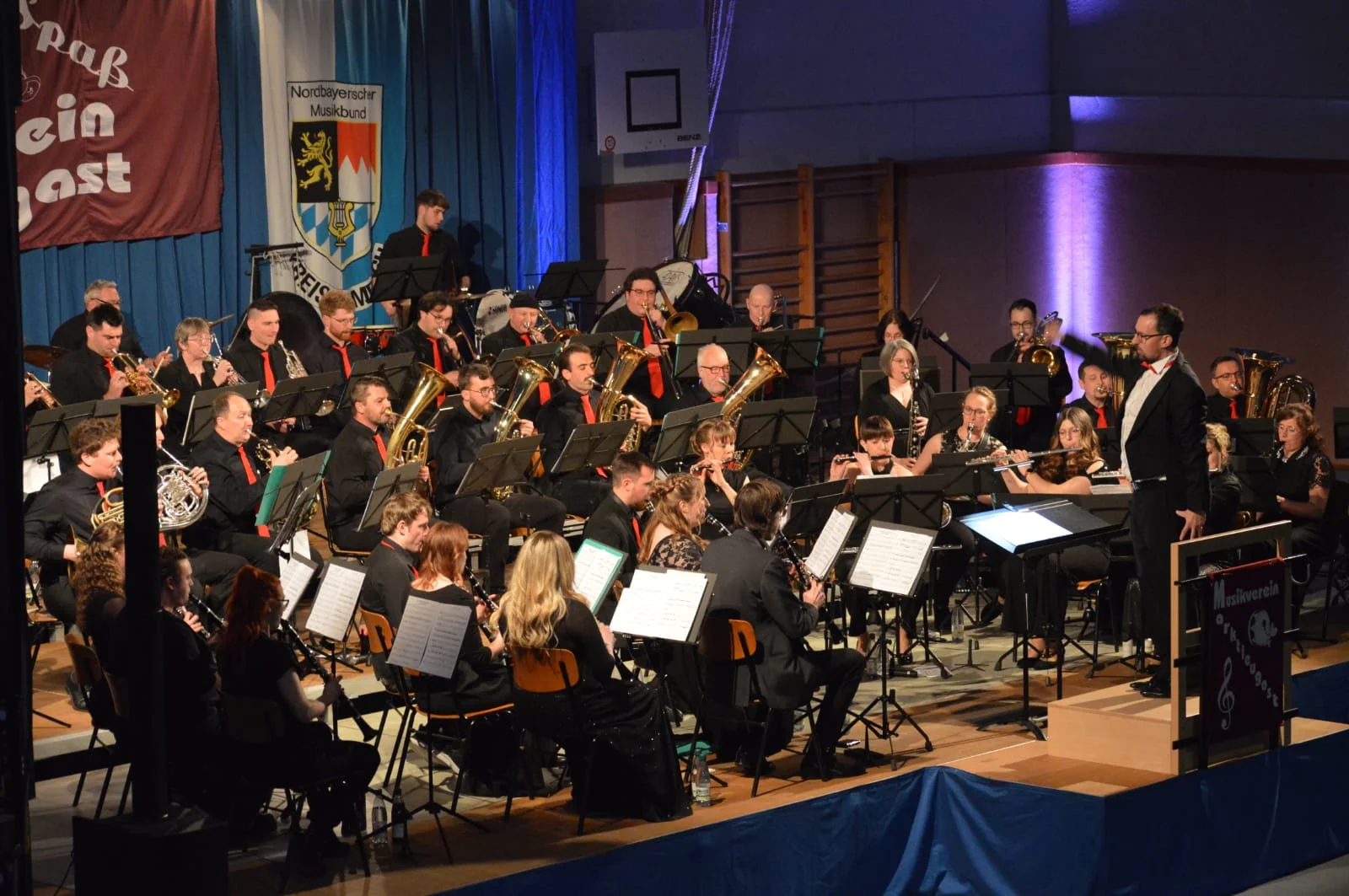 Alle Musikerinnen und Musiker in schwarzer Kleidung sitzen auf der Bühne und tragen hochkonzentriert ein Musikstück vor. Die Männer tragen alle eine rote Krawatte. Der Dirigent trägt einen edlen Frack und dirigiert energisch.