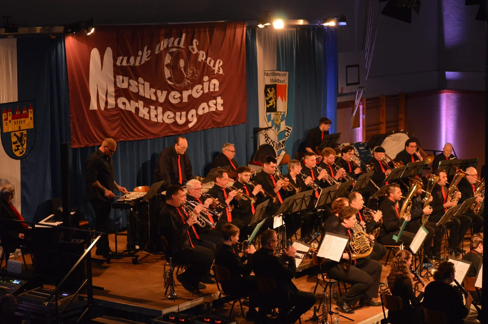 Alle Musiker sitzen aufgereiht nebeneinander auf der Bühne und halten ihr Instrument vor sich am Mund. In der hinteren Reihe stehen einige Schlagzeuger vor dem Schlagwerkinstrumenten. Dahinter weht das große Logo des Musikvereins und die Fahnen von Marktleugast und des Nordbayerischen Musikbundes.