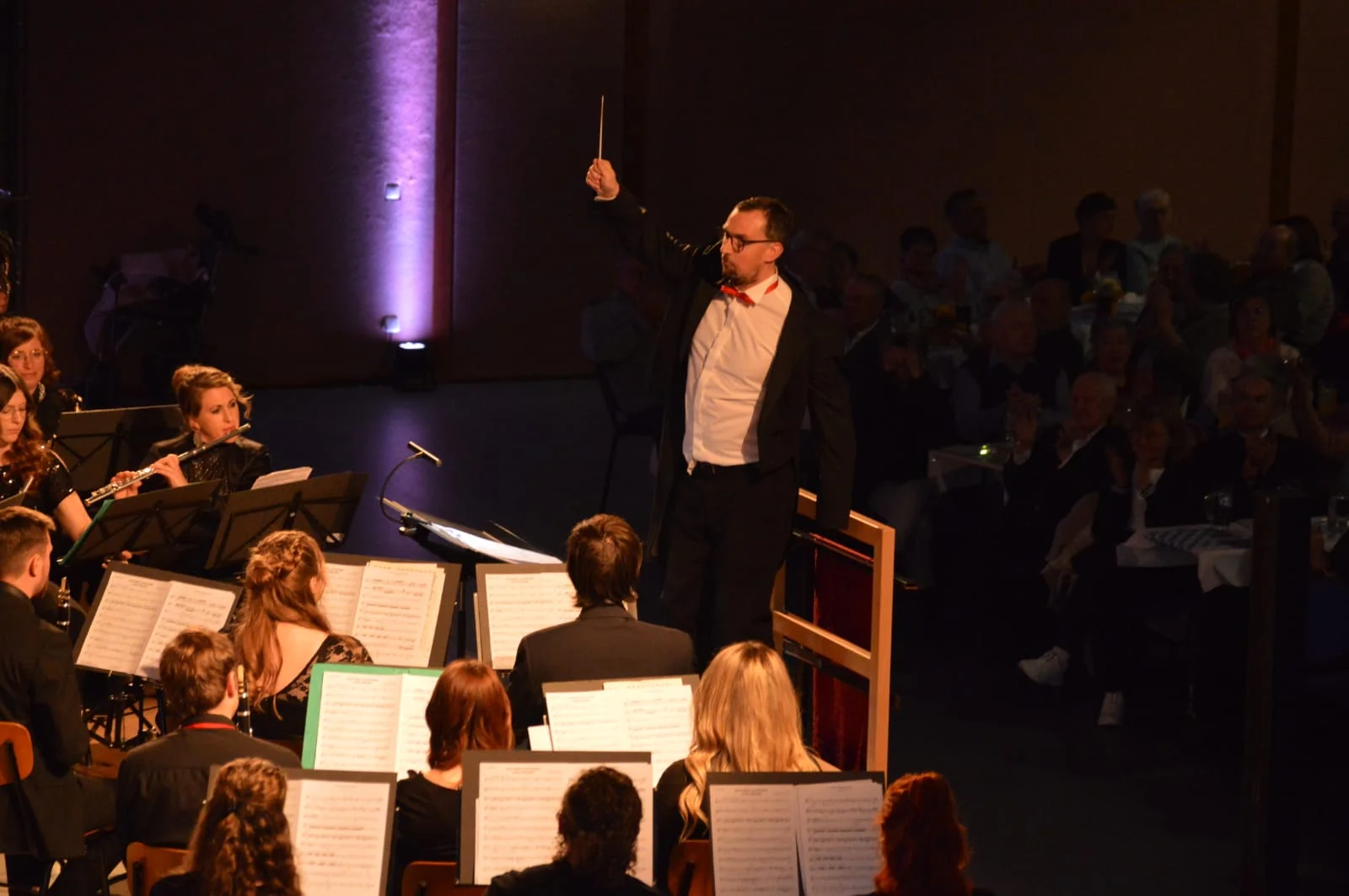 Der Dirigent hält den Dirigentenstab hoch in die Luft. Einige Musiker sind von hinten zu sehen. Sie sehen alle in die Noten die vor Ihnen auf Notenständer liegen.