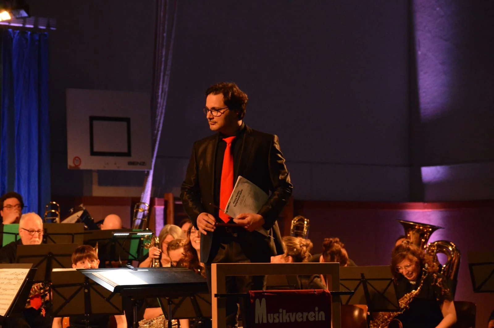 Der zweite Dirigent Dr. Matthias Gebauer steht vor dem Orchester und hält Noten und Dirigirstab in der Hand. Im Hintergrund lächeln die Musikerinnen und Musiker.