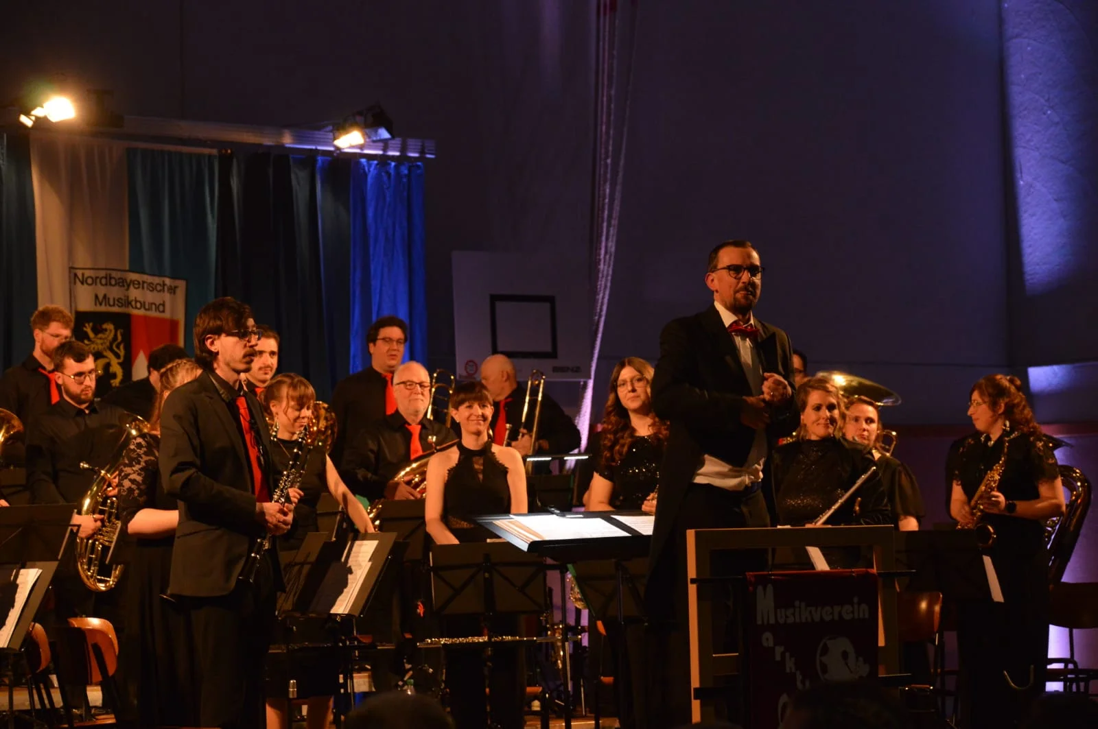 Nach einem Stück stehen alle Musiker und lächeln in den Zuschauerbereich. Auch der Dirigent ist den Zuschauern zugewandt.