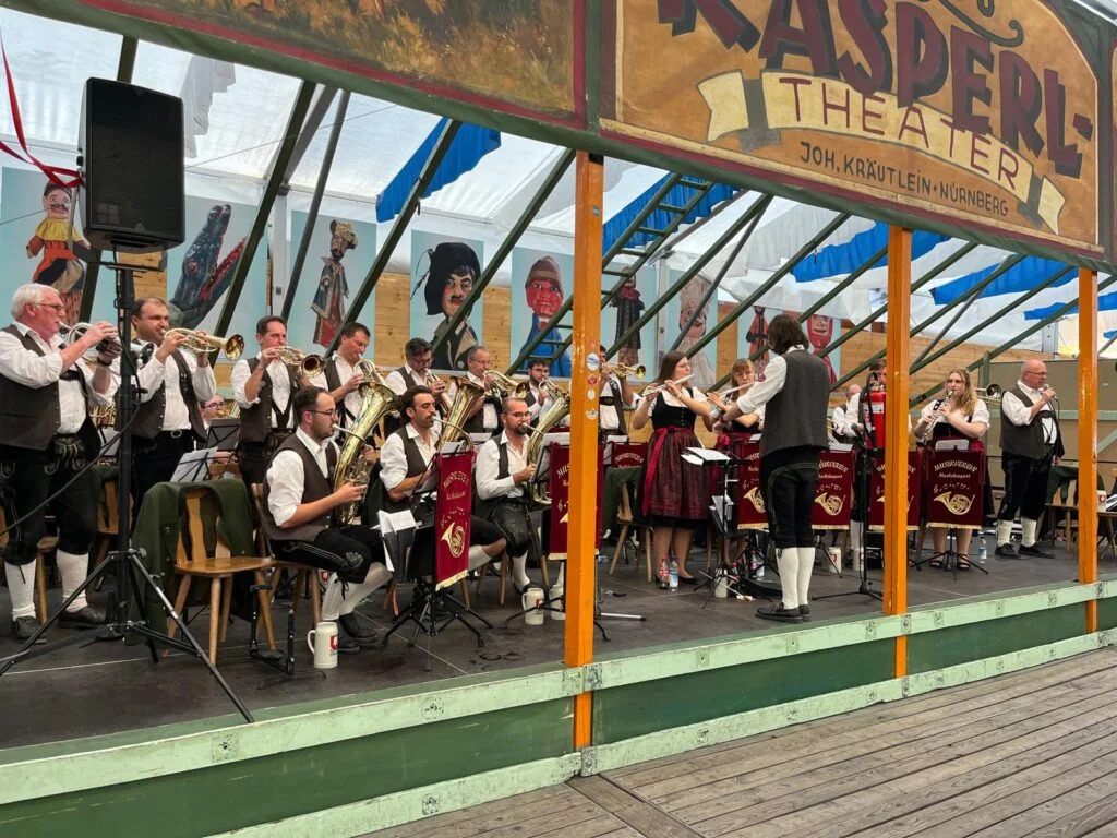 Die rund 30 Musikerinnen und Musiker vom Musikverein auf der Bühne beim Oktoberfest 2026. Die Trompeten und Klarinetten stehen. Dazu wird gesungen. Die Bühne steht im Museumszelt auf der Oidn Wiesn. Oben ist der Aufdruck Kasperl-Theater zu lesen.
